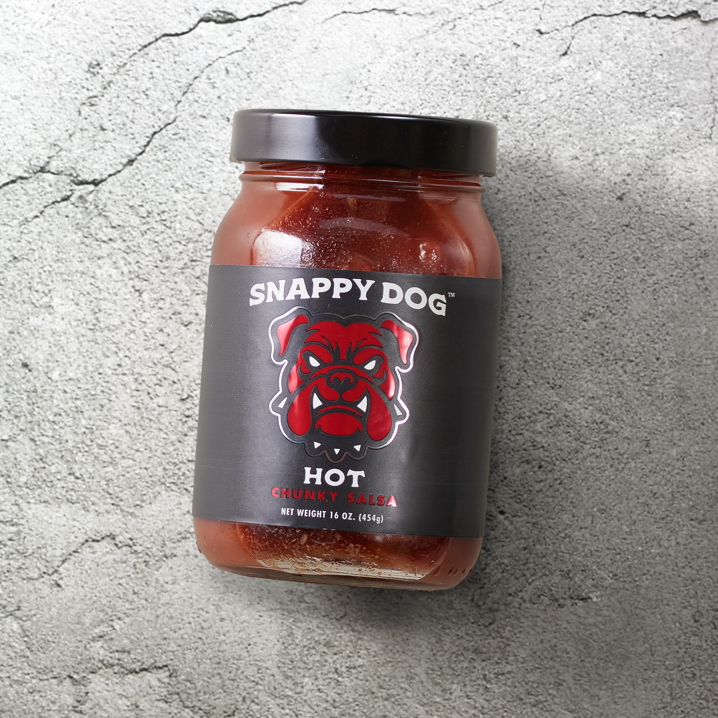 Jar of Snappy Dog Hot chunky salsa, with black label, that has metallic red dog head of snappy dog logo.