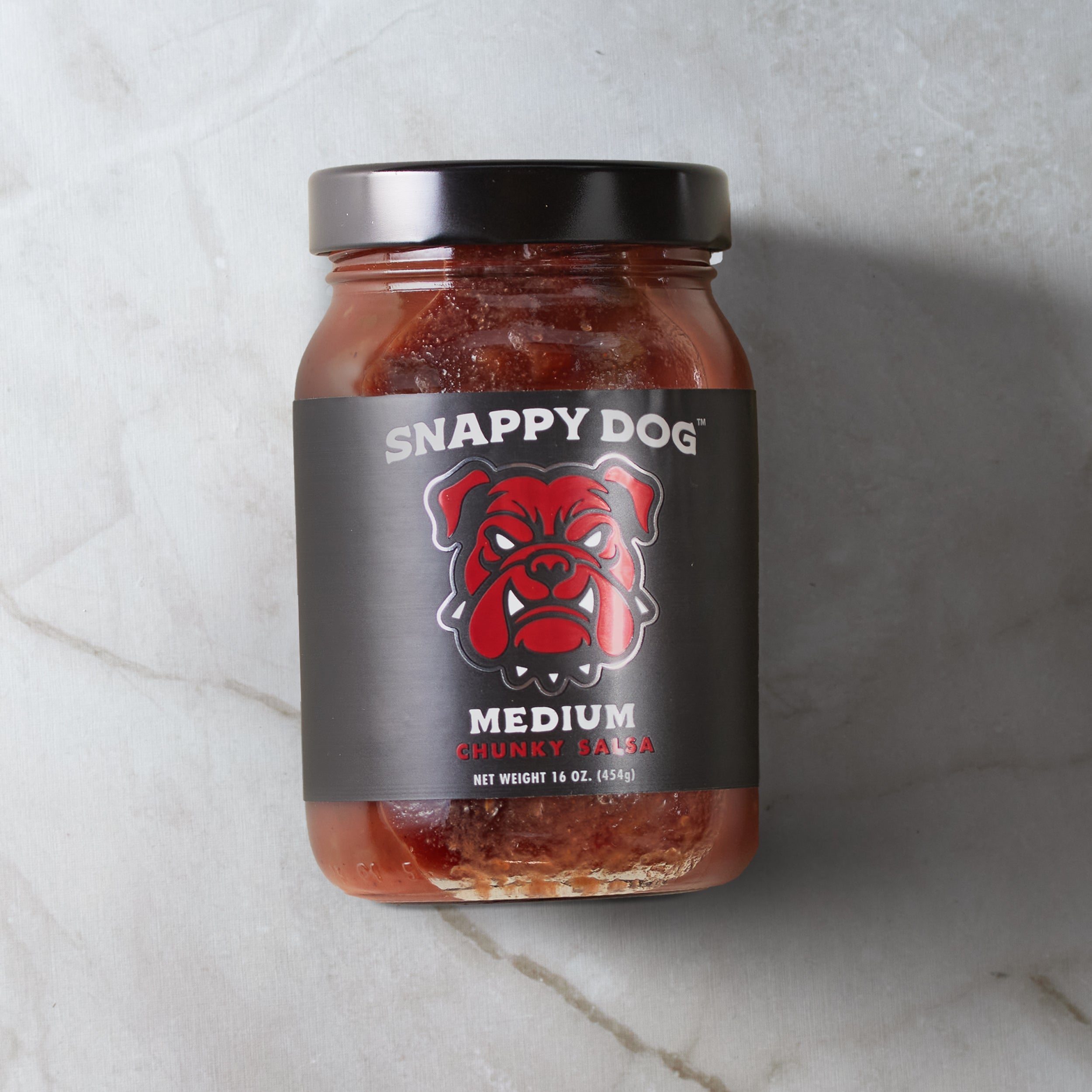 Jar of Snappy Dog Medium chunky salsa, with black label, that has metallic red dog head of snappy dog logo.