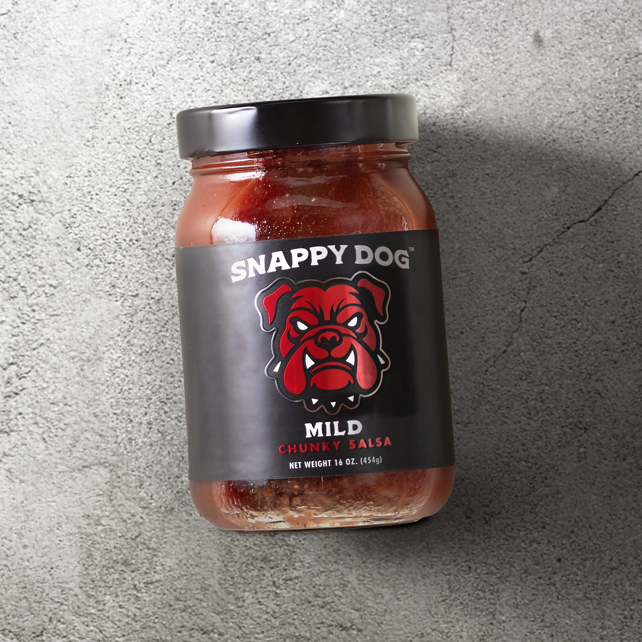 Jar of Snappy Dog Mild chunky salsa, with black label, that has metallic red dog head of snappy dog logo.